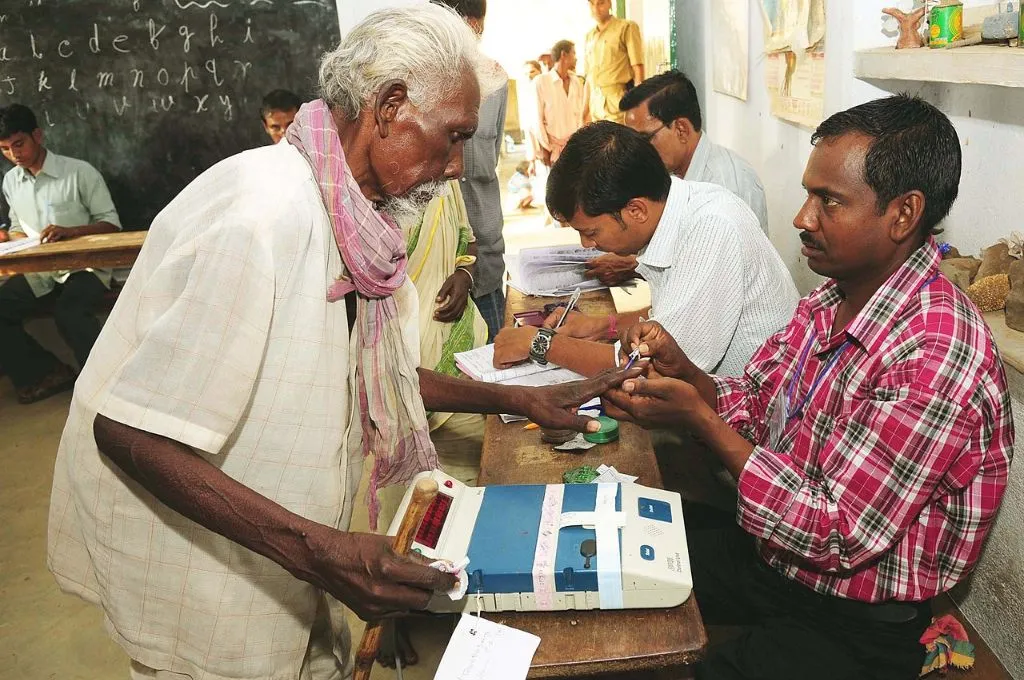 The image shows an elderly male voter inside a polling booth station in a village. He stands in front of a table as a polling booth officer applies blue ink to his index finger.--local self-governance