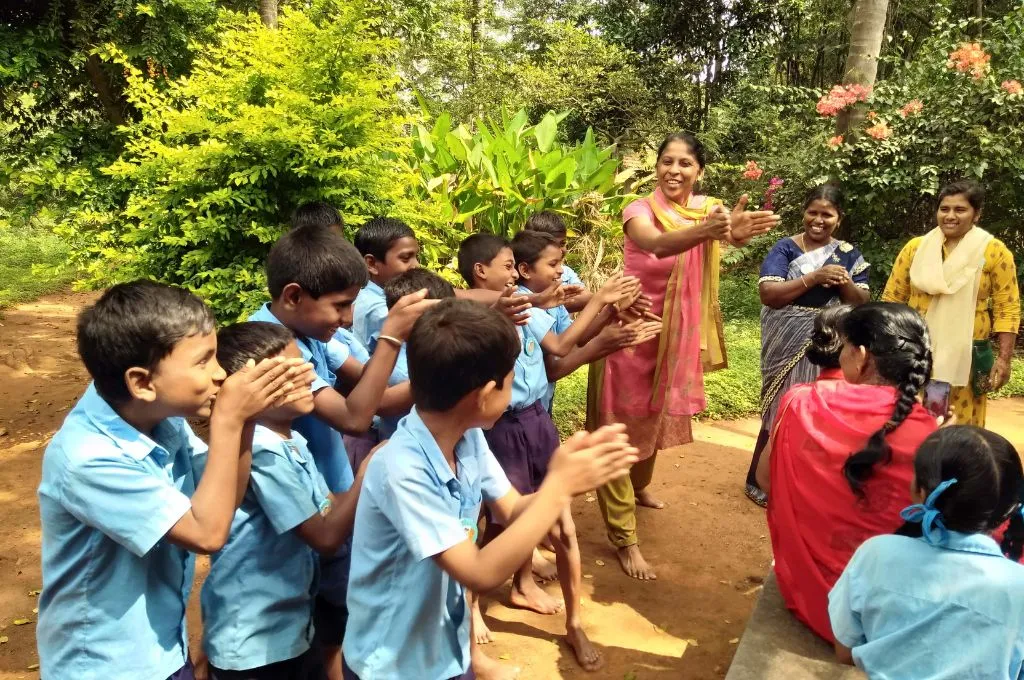 Young schoolchildren in light blue uniforms stand on a dirt path and clap together as a woman in a pink and yellow salwar kameez leads an activity. Other women stand nearby and smile, while green trees and flowering plants frame the lively outdoor scene--environmental studies (EVS)