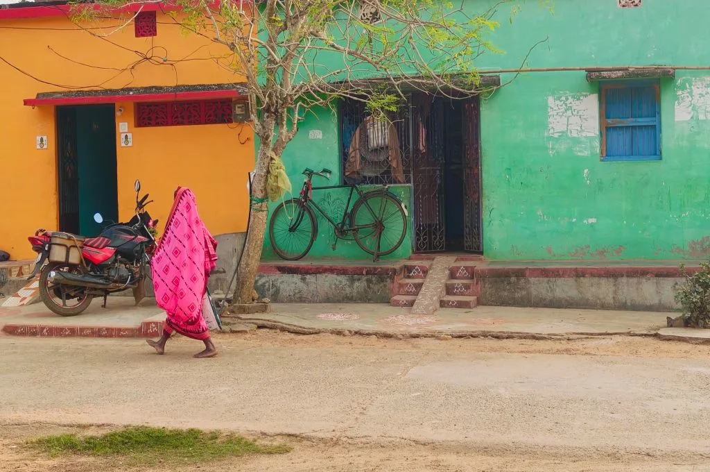 A person wrapped in a bright pink sari walks along a quiet street past colorful houses with green and yellow walls, where a bicycle leans against a doorway and a motorcycle is parked nearby--union budget india