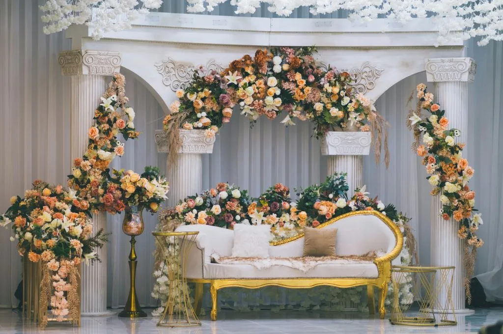 A wedding stage setup featuring white classical columns and arches decorated with peach, cream, and white floral arrangements. A white and gold vintage-style loveseat sits at the center, surrounded by floral garlands and golden tables--nonprofit humour