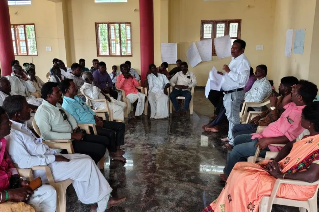 a man standing up and addressing a group of people seated in a semi circle inside a hall--community forest rights