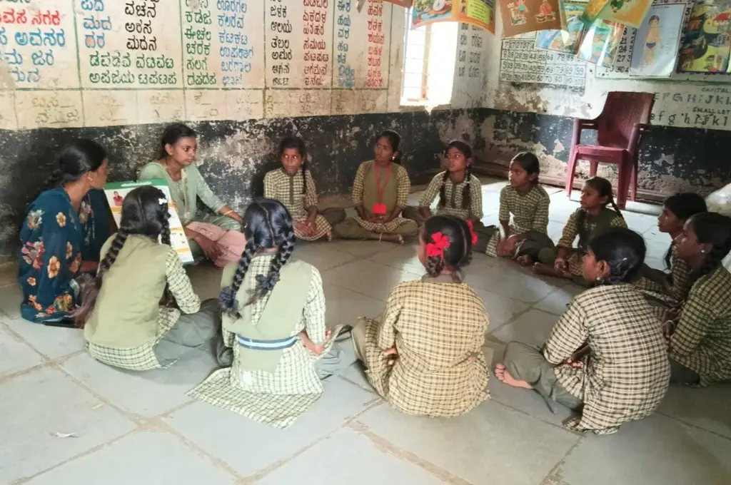 young girls in school uniforms sitting on the classroom floor in a circle and being addressed to by two women--community organiser