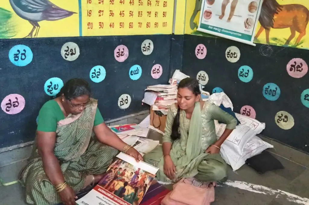 two women sitting on the floor in a classroom, with numbers and the kannada alphabet painted on the walls behind them--community organiser