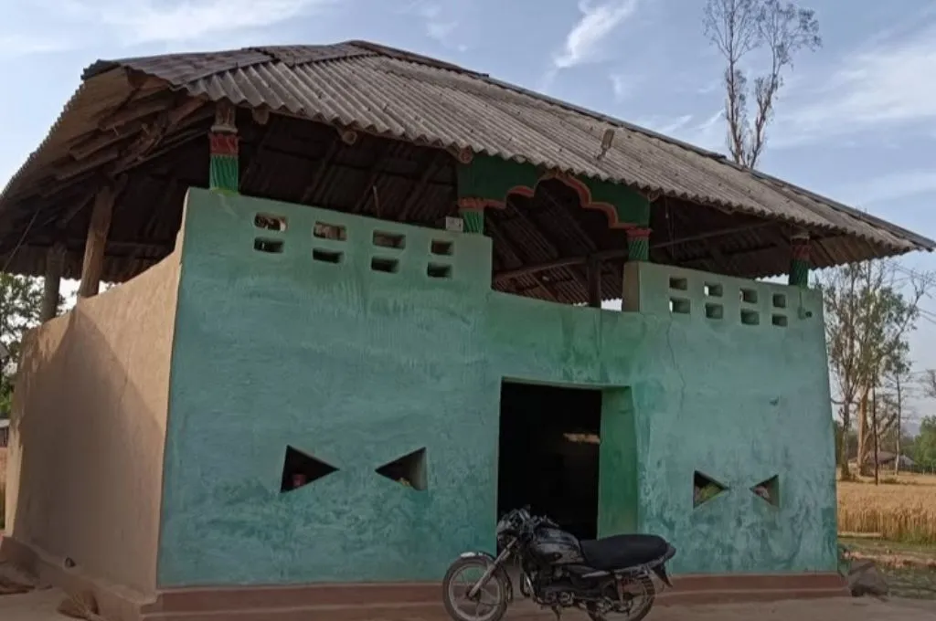 A small mud house with green painted walls and a corrugated metal roof, featuring triangular ventilation openings and an open doorway. A motorcycle is parked in front, and dry vegetation and a few trees are visible in the background under a partly cloudy sky--Van Gujjars