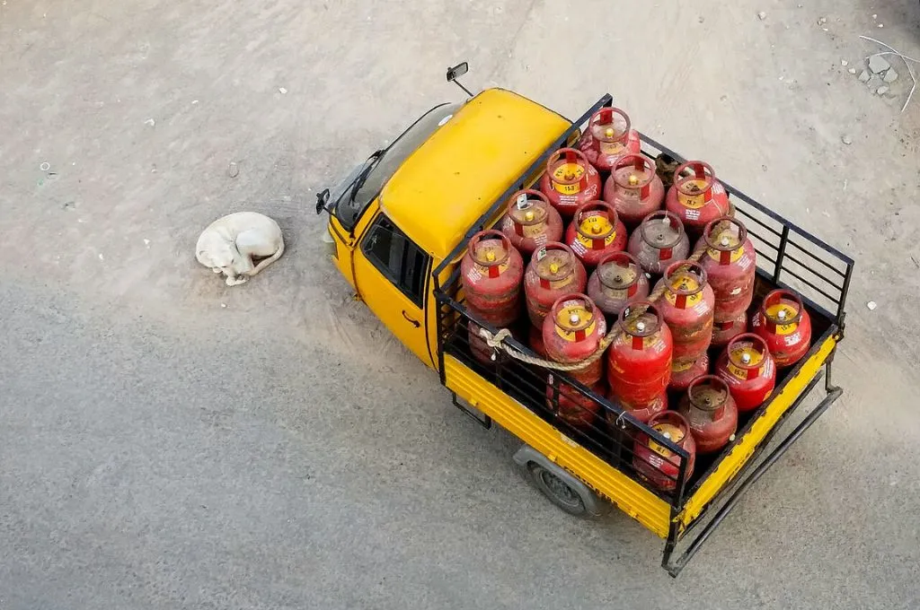An overhead view of a small yellow truck loaded with multiple red LPG gas cylinders parked on a dusty ground. A white dog lies curled up beside the truck, resting on the bare surface nearby--LPG shortage in India