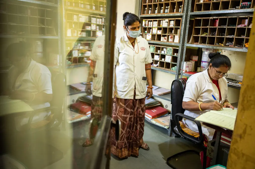two female healthcare workers in a pharmacy, with one writing in a register while the other looks on--public health India