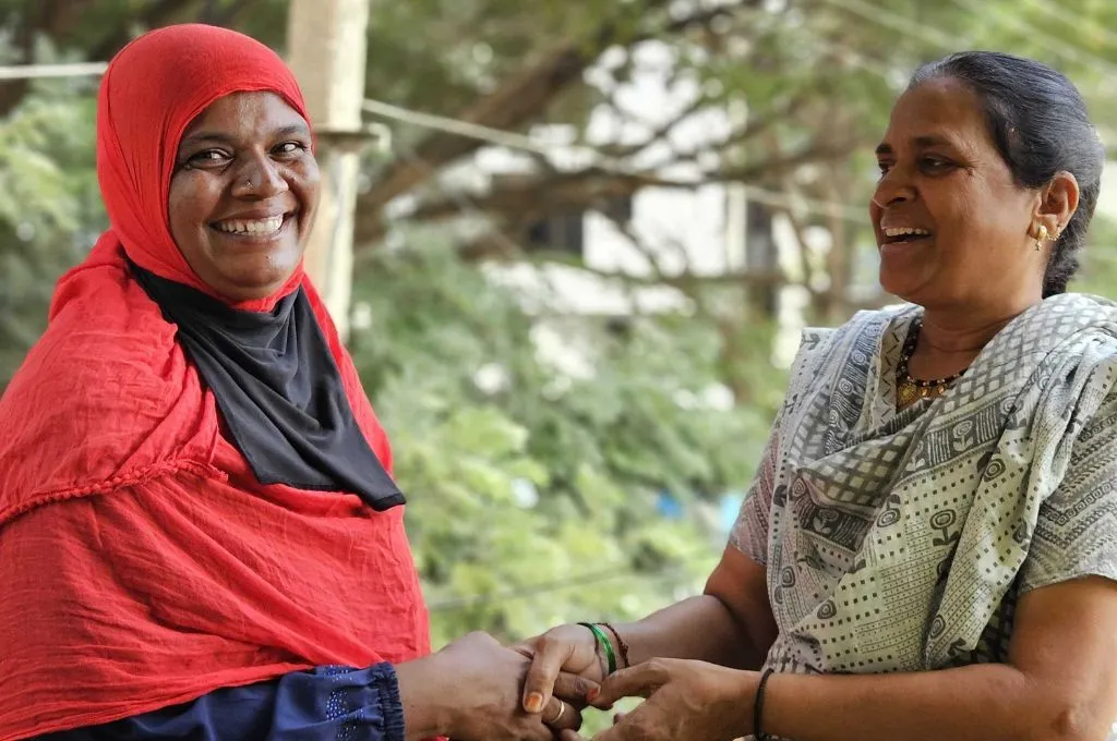 The image features the authors, Shakeela Banu and Nageena Banu. They are standing outdoors smiling warmly at each other while holding hands. Shakeela is wearing a bright red headscarf with a black inner layer, and Nageena is wearing a light-colored and patterned suit and dupatta . The background shows greenery and a blurred building--Beedi industry