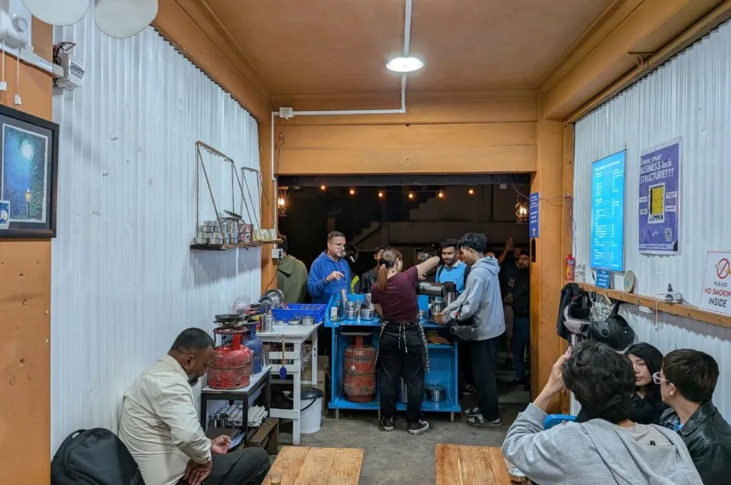 a few people seated inside a tea shop, with the owner standing behind the counter and serving a few other customers standing around her--sustainable business