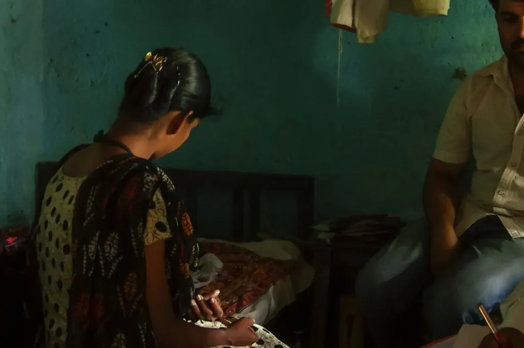 The image is set inside a home. A young women is sitting at the edge of a bed with her back turned to the camera. She is holding the hand of a person who is lying down on the bed, out of the frame.--palliative care