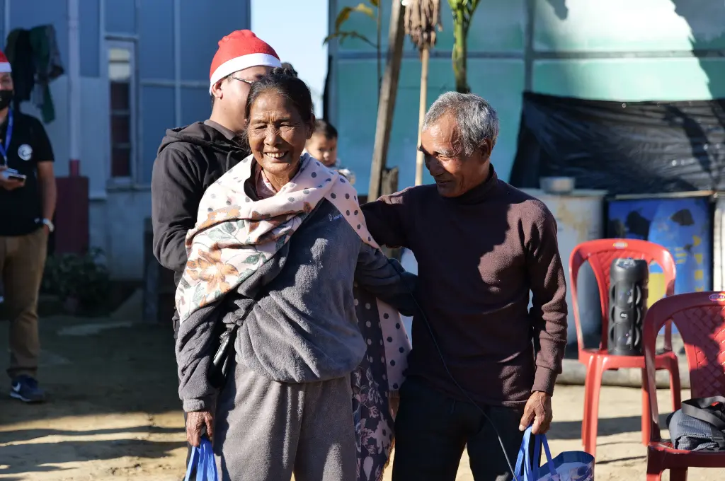 an elderly woman smiling with an arm around an elderly man who is looking at her; in the background is a baby and two men with santa hats--crowdfunding in Mizoram
