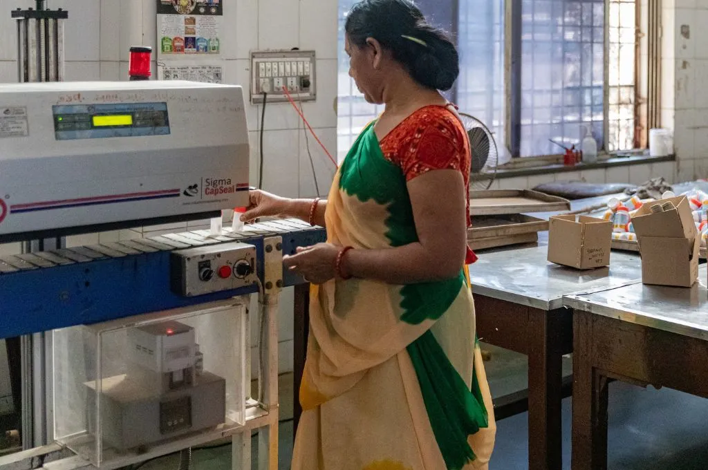 A woman in a sari operates a capsule sealing machine in a small laboratory or production room, with boxes and equipment arranged on nearby tables--non communicable diseases india