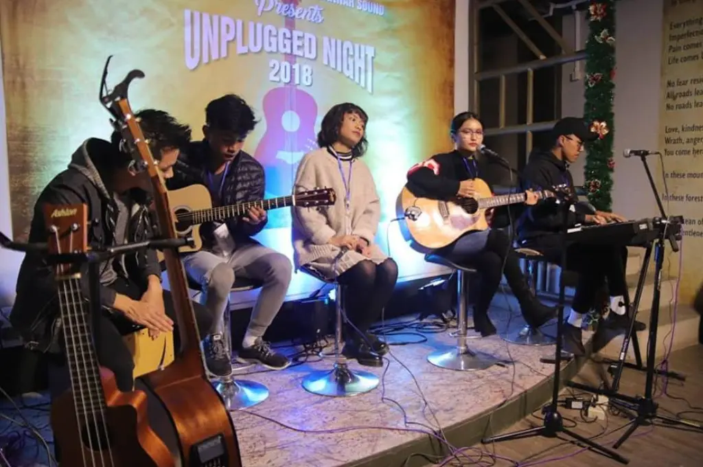 a band, with some members holding guitars, on a stage in fromt of a screen that says "Unplugged Night 2018"--Mizoram nonprofits