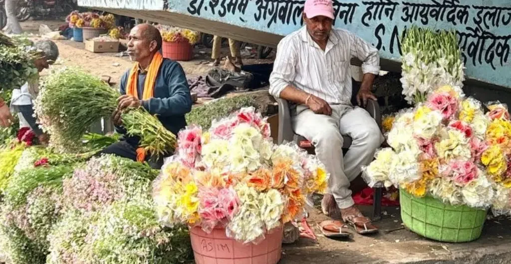 The image shows two men sitting on a footpath. They are selling flowers of different colours which are bundled together in plastic buckets and arranged in heaps on the ground.--floriculture