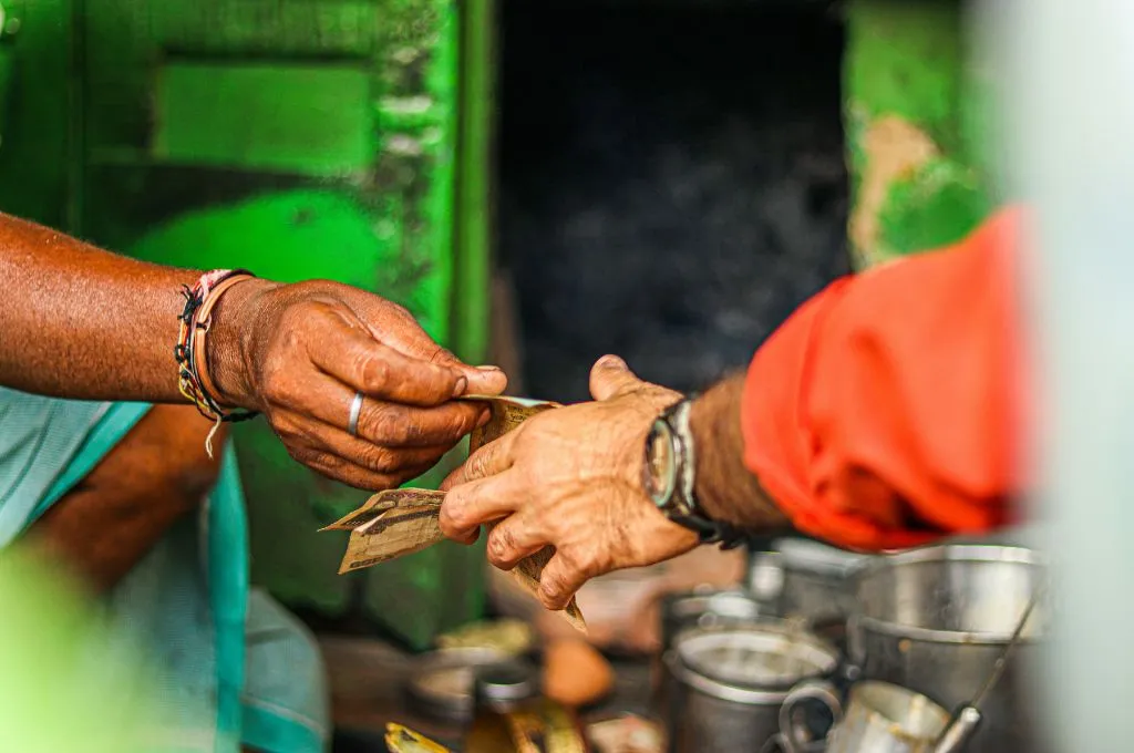 Close-up of two people exchanging paper currency by hand at a small street-side shop, with metal cups and containers blurred in the background--funders