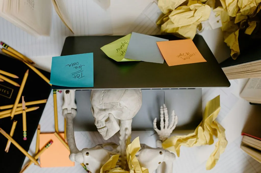 Overhead view of a skeleton figure lying at a cluttered desk with a laptop on its chest, surrounded by sticky notes, pencils, crumpled paper, and books--nonprofit humour