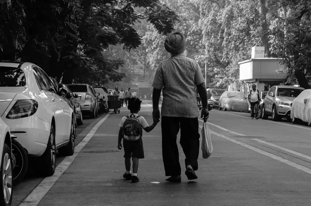 a man holding the hand of a small child with a schoolbag, walking down a street with their backs towards the camera--cost of schooling