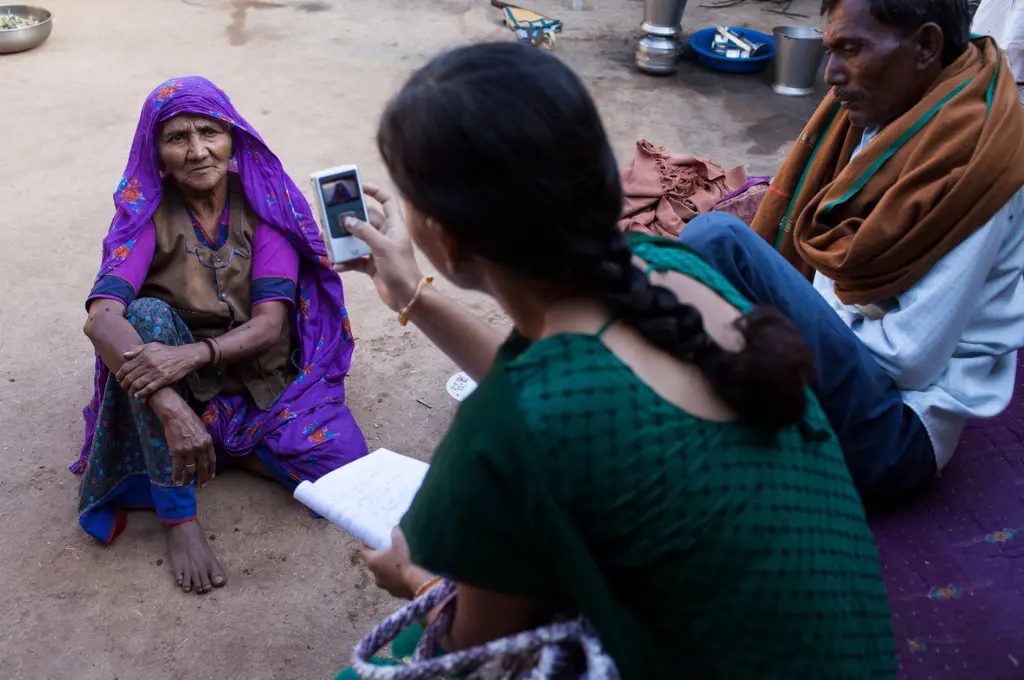 an elderly woman sitting on the ground speaks into a camera being held by a female journalist--data collection