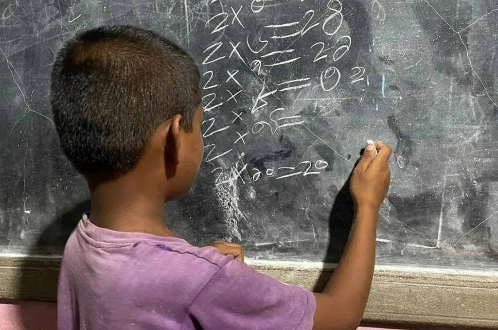 A child in purple tshirt writes multiplication problems with chalk on a worn blackboard in a classroom--foundational education
