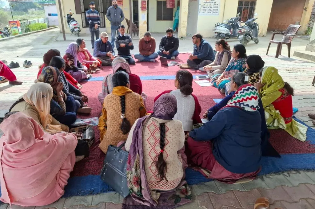 A group of people sit in a circle on a mat outside a building, participating in a group discussion or training session--digital tools