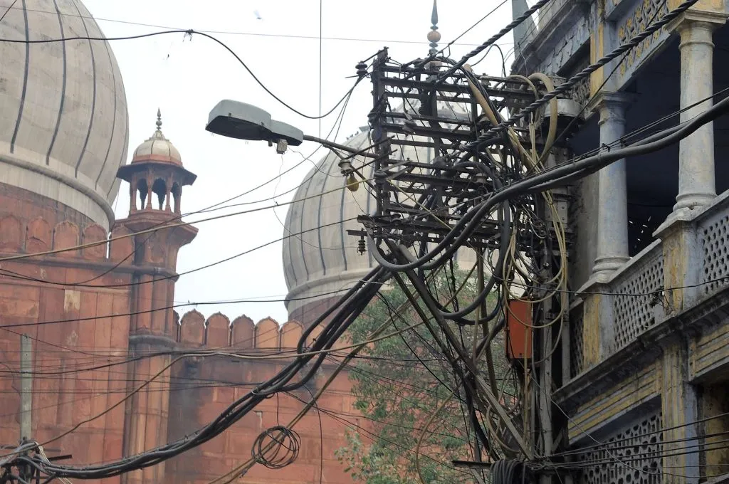 A cluster of electrical wires and utility cables hangs from a pole beside a monument. Large domes and red sandstone walls in the background--nonprofit AI