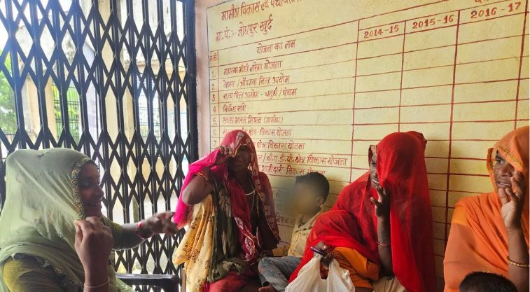 a group of woman talking to each other; on one side is an iron sliding door and behind them is a wall with a chart painted on it--MGNREGA