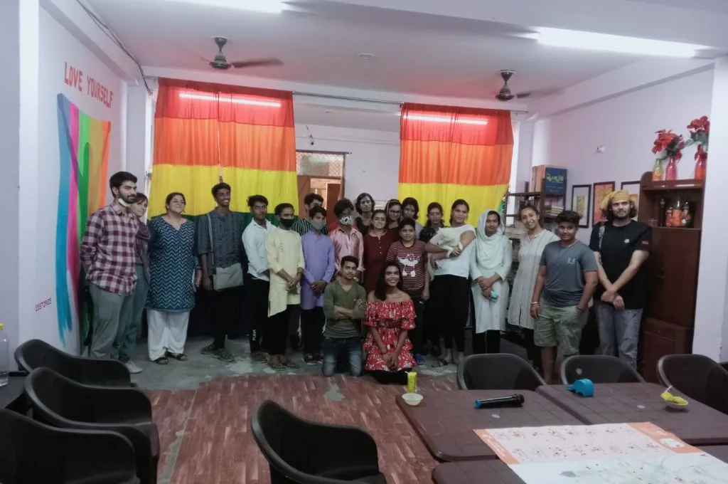 A large group of people in a room posing for a group photo; one of the rooms in the wall has a pride flag over which is written 