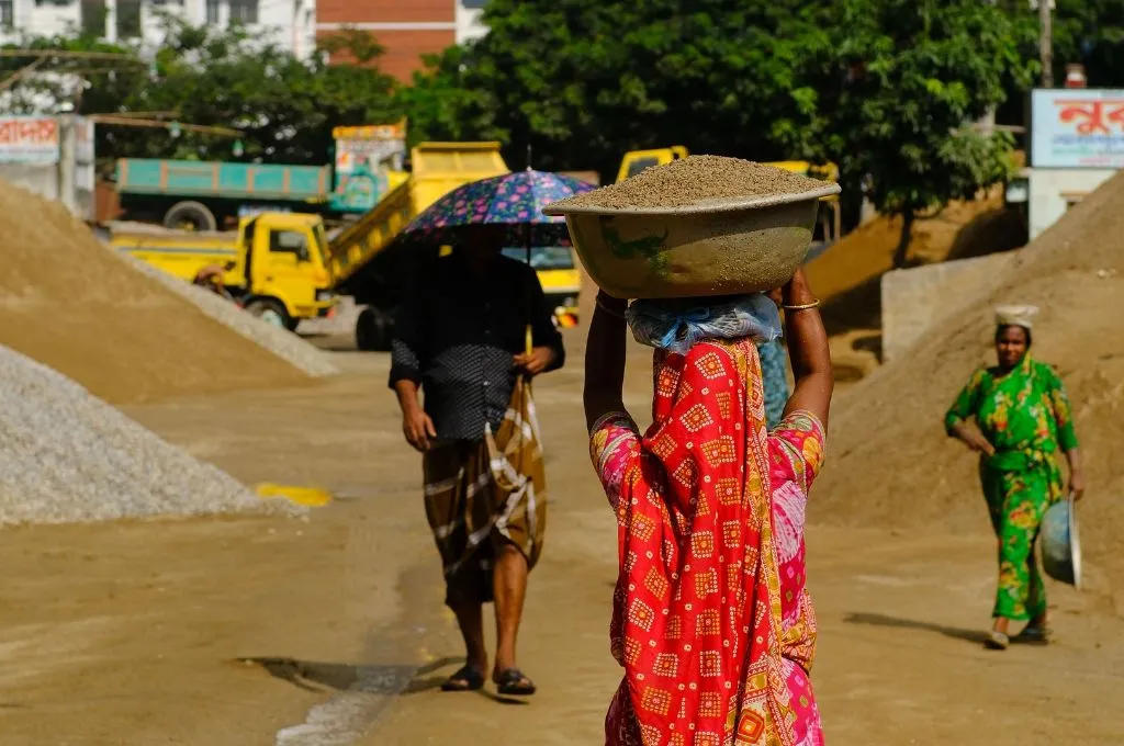 A construction worker in sari carries a large basin of sand on her head at a construction site, with piles of gravel, trucks, and other workers visible in the background--Heat budget