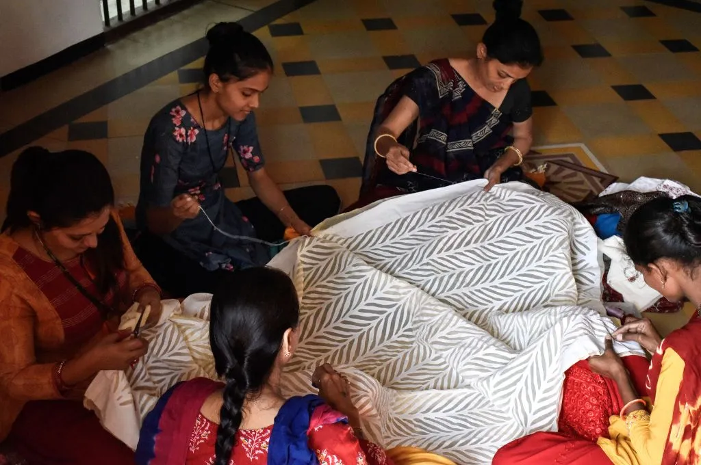 Five women seated on the floor work together, hand-stitching a large white fabric with a gray leaf pattern in an indoor space--heat and humidity