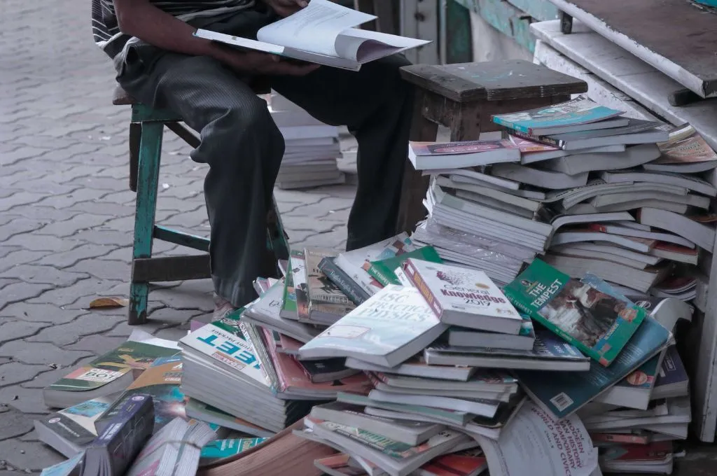 books stacked roughly on the footpath; sitting next to the stack is a person looking through a book--preparatory spaces