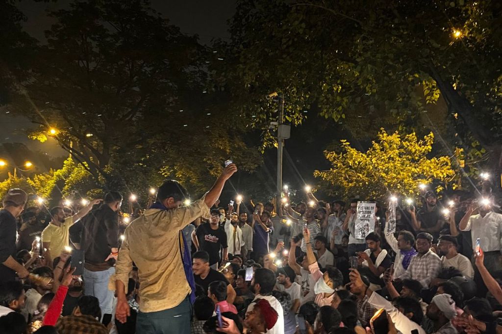 A crowd gathers outdoors at night, holding up lit mobile phones during a peaceful demonstration beneath trees--protest in india