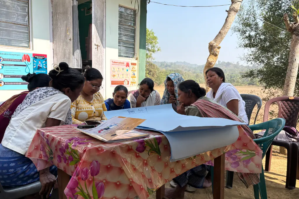 a group of women sitting around a table with charts and a booklet in it huddled over something--Meghalaya health data