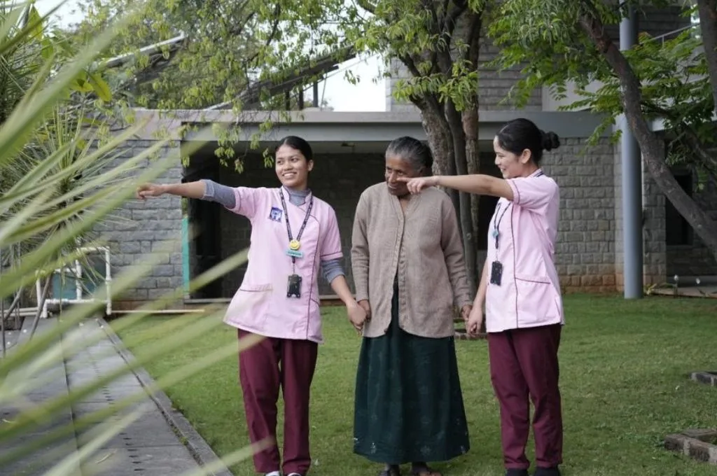 two nurses walking with an elderly woman in a garden surrounded by walls--hospice facilities