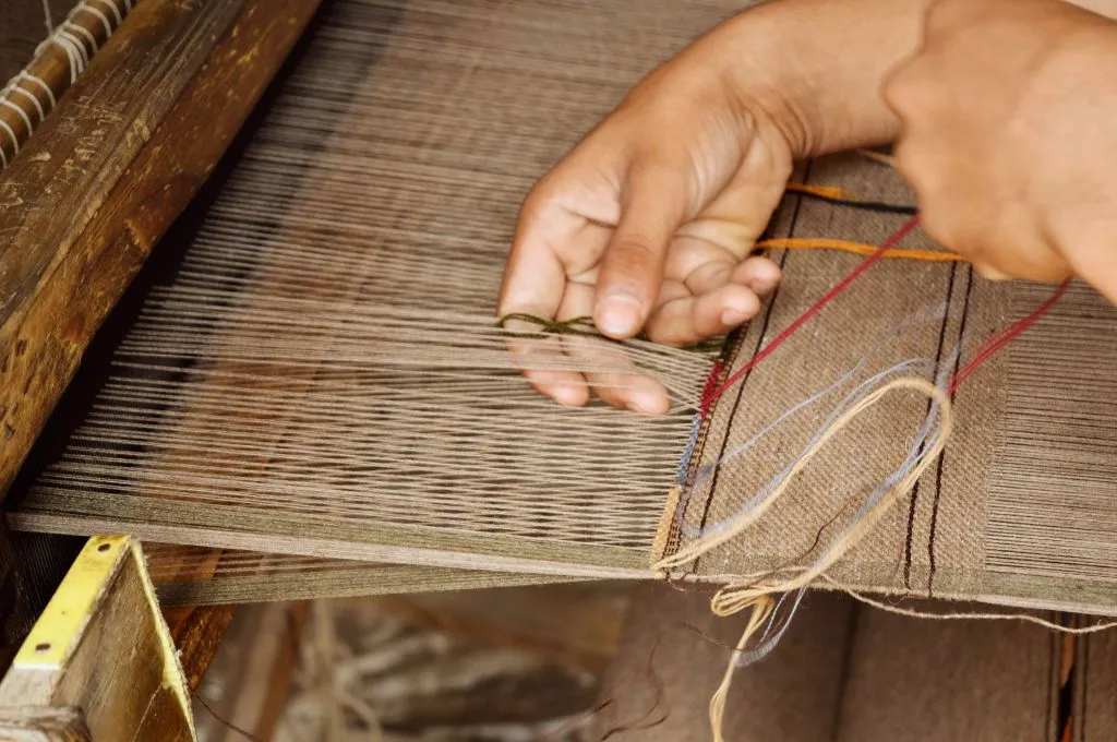 close-up of a person weaving fabric on a handloom, guiding threads by hand as yarns run through the loom._CSR india