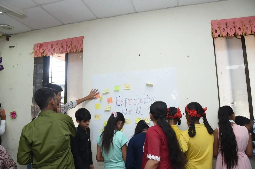 a group of children standing in front of a white wall that has a chart titled "Expectations Wall' pasted on it--unconference