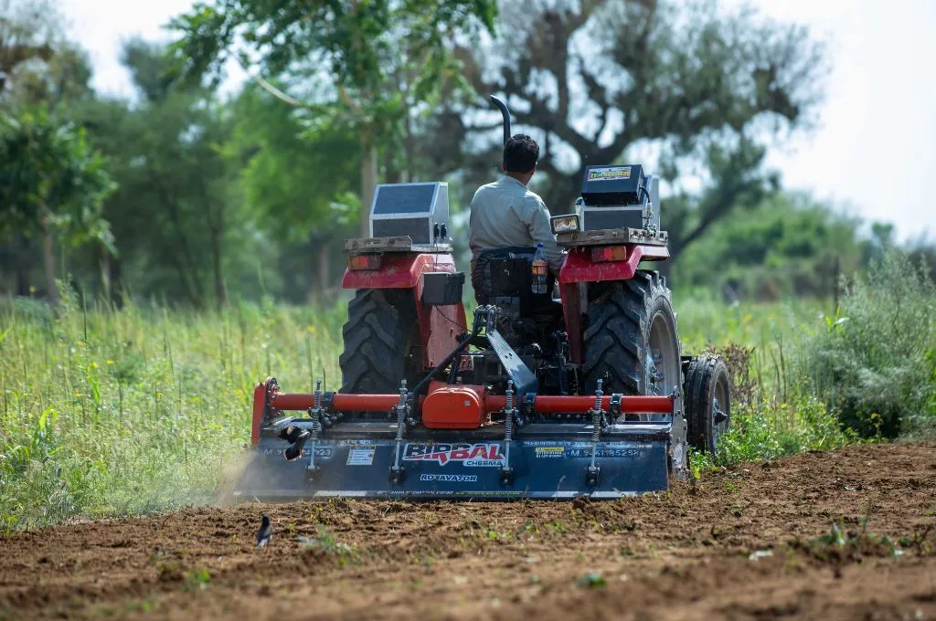 A farmer on a tractor tills dry soil in a field with trees and grass in the background--carbon farming