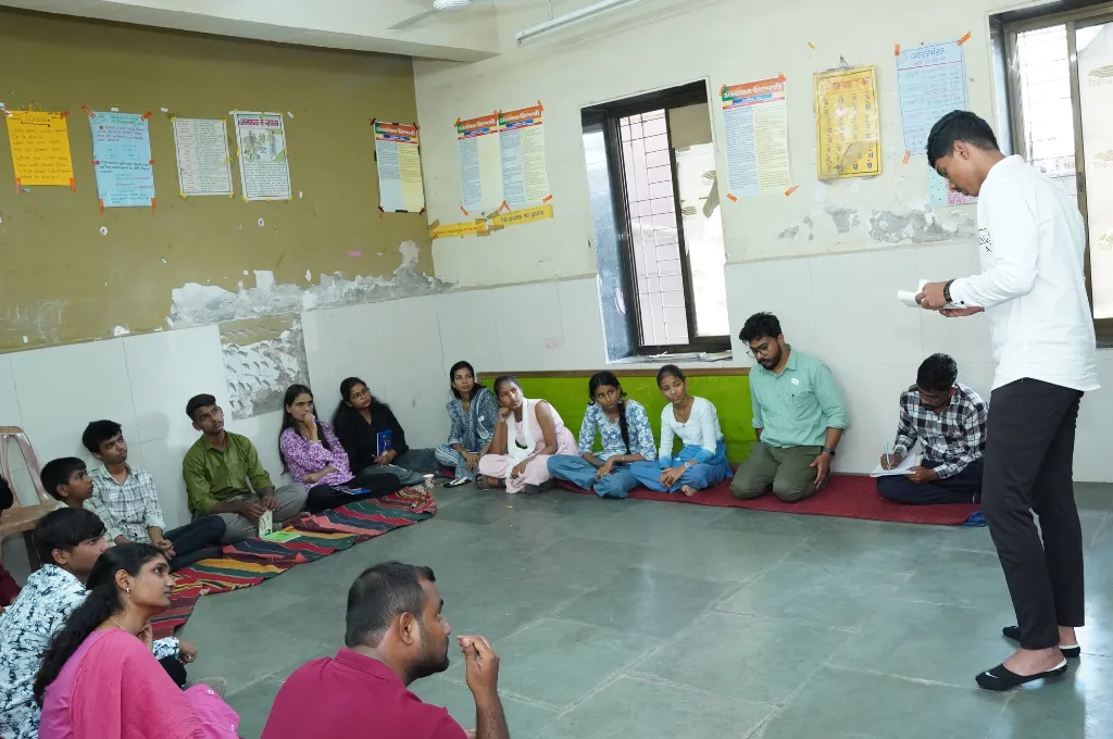 a group of people sitting on the floor along the walls of a classroom listening to a young man read something off of a sheet of paper--