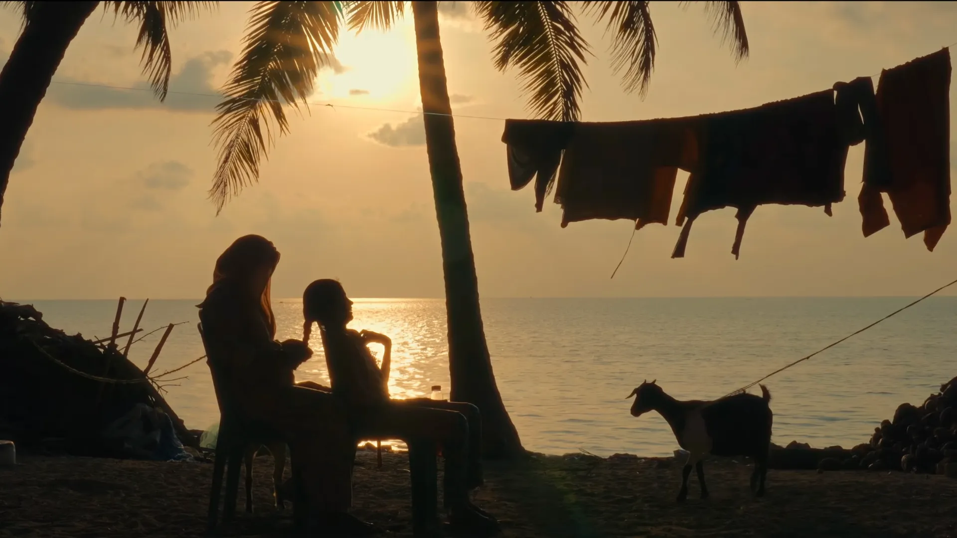 a woman braiding a young girl's hair on a beach as the sun sets. to their right is laundry drying on a wire and a goat that's been tied up to a palm tree--community engagement in Lakshadweep