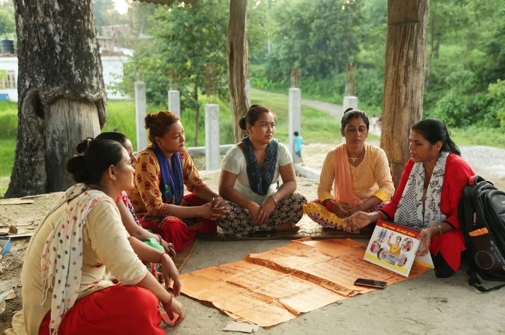 Five women sit together outdoors under a large tree, discussing materials spread on the ground, with green landscape visible behind them--gender-based violence