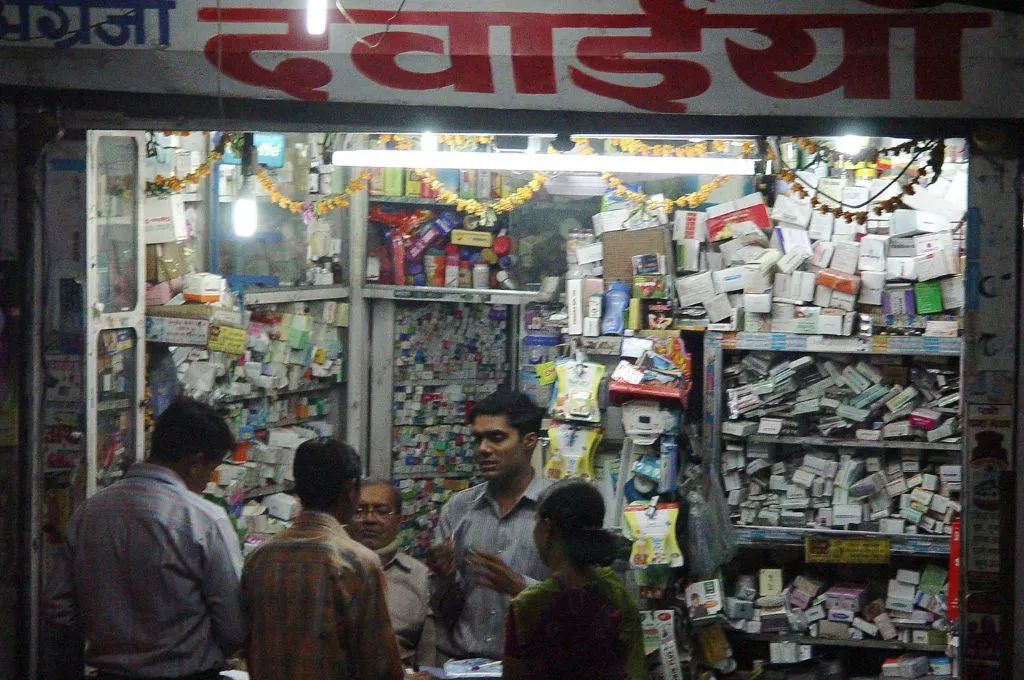 A crowded pharmacy shop with shelves and counters packed with medicine boxes, while customers speak with the shopkeepers--contraceptive packaging