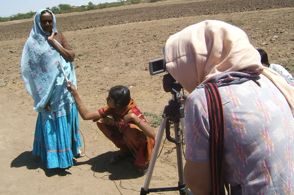 a woman standing in an empty field speaking into a camera being manned by another woman while a young girl squatting on the floor holds up a mike--mental health