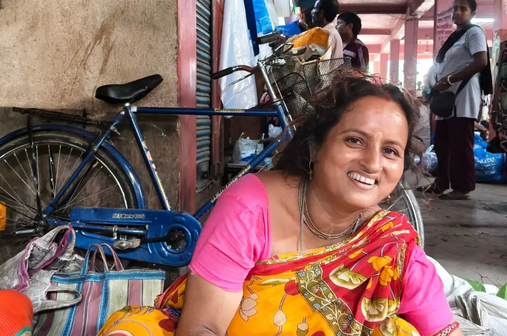 a woman in a bright saree smiling into the camera. In the background is a bicycle leaning against the wall and a corridor full of people--SIR