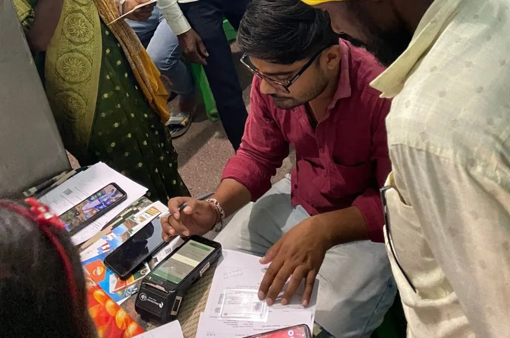 a man in a read shirt and black-rimmed glasses sits on the floor and verifies government IDs against data on a small digital device as people stand around him--fertilisers