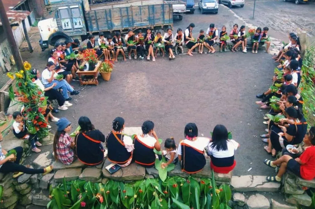 The image features young women and girls seated in an an open area. They are dressed in traditional attire and eating food served in plates made from leaves._KhuKhu Nye