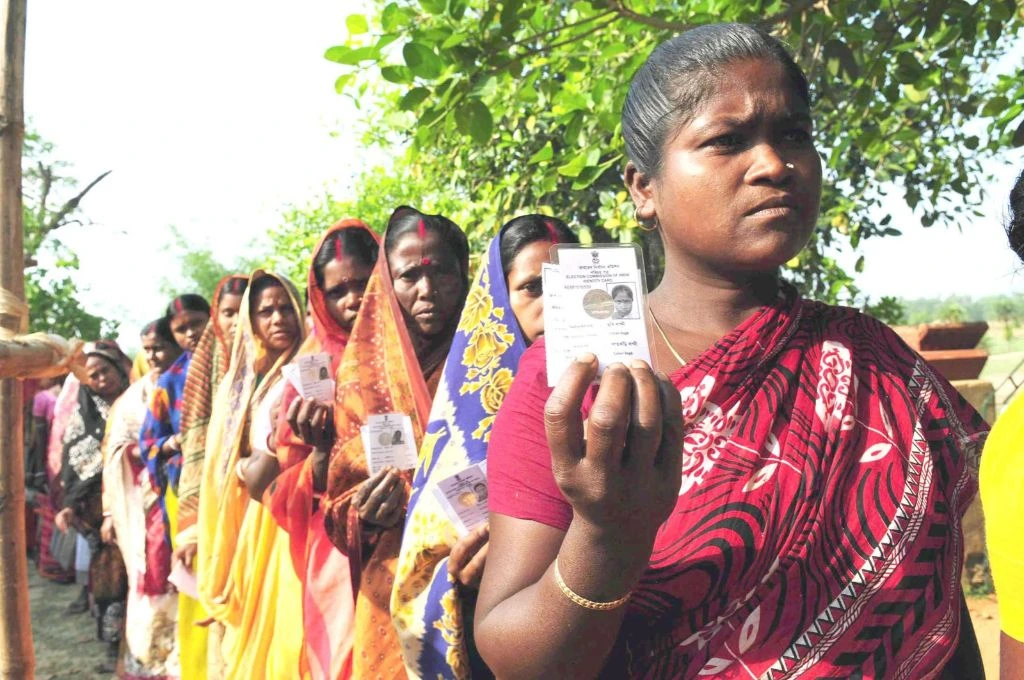 The image features a group of women standing in a single file. Each is holding up her voter ID card and looking ahead.--SIR